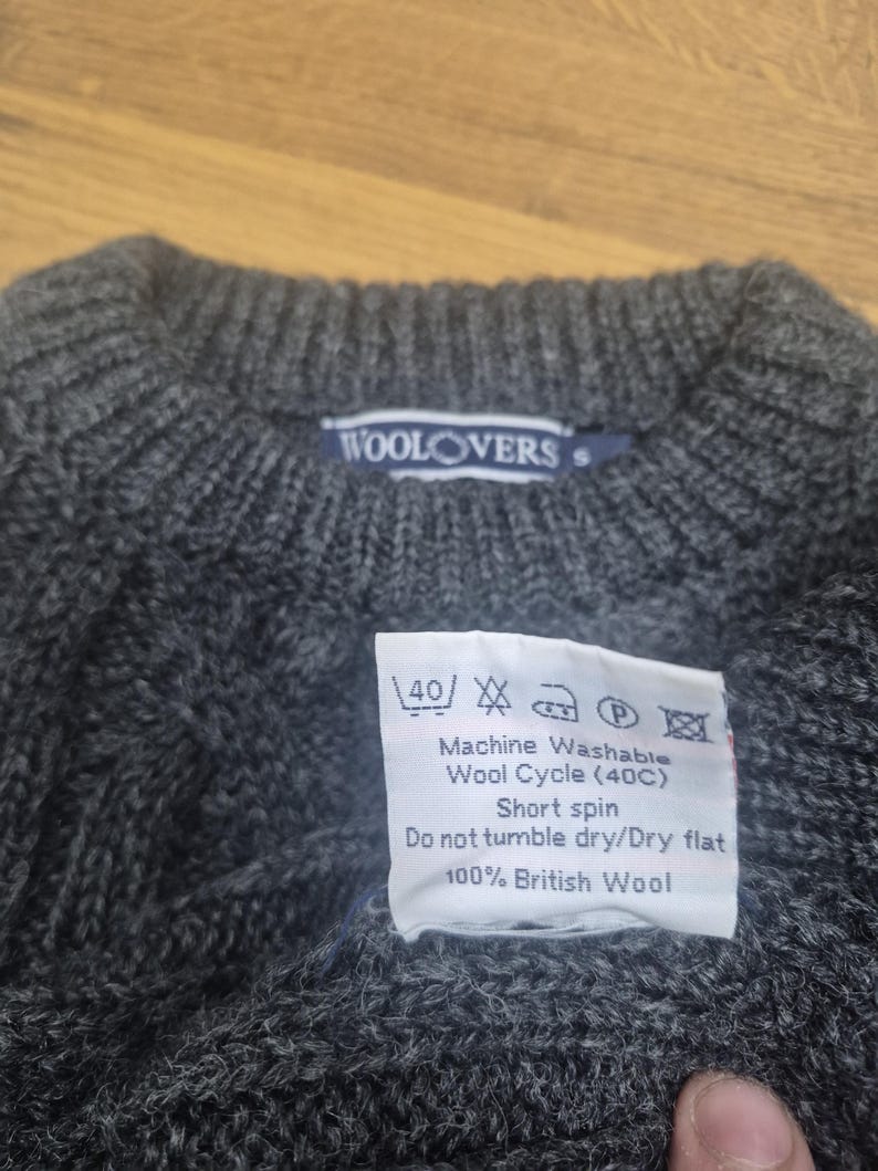 May include: A dark gray wool sweater with a crew neck. The label reads "WOOLOVERS". A tag indicates the sweater is machine washable at 40 degrees Celsius and made of 100% British wool.