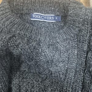 May include: A close-up of a gray cable-knit sweater. The sweater has a ribbed collar and a small blue tag that reads "WOOLOVERS" and the size "S". The knit pattern is visible throughout the sweater.