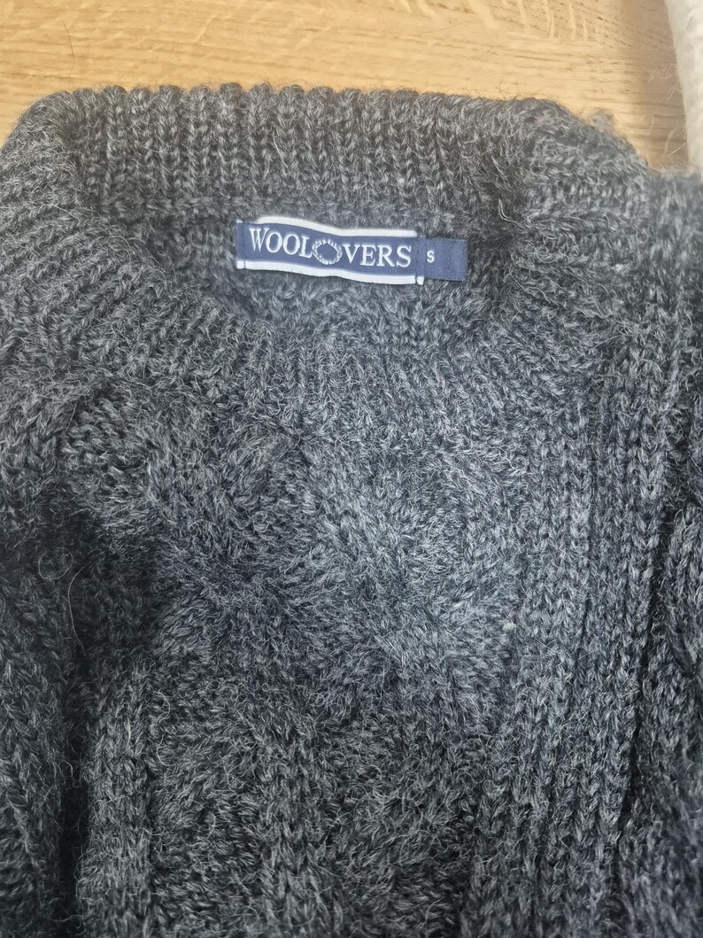 May include: A close-up of a gray cable-knit sweater. The sweater has a ribbed collar and a small blue tag that reads "WOOLOVERS" and the size "S". The knit pattern is visible throughout the sweater.