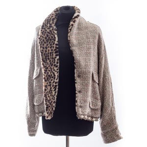 Könnte beinhalten: Tweedjacke mit Leopardenmuster-Kragen und -Besatz. Die Jacke hat zwei Vordertaschen und einen Knopfverschluss. Der Tweed ist eine Mischung aus braunen, grauen und roten Fäden. Die Jacke wird auf einer schwarzen Schaufensterpuppe präsentiert.