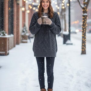 May include: A woman wearing a gray cable-knit sweater, black leggings, a brown knit hat, and brown boots stands in the snow. She holds a wooden mug with a warm beverage. The background features a snowy street with string lights.