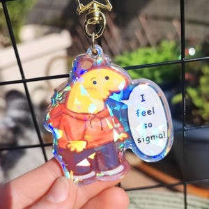 Puede incluir: Un llavero holográfico transparente con un personaje de dibujos animados con una sudadera con capucha naranja y pantalones azules. El personaje tiene un bocadillo que dice "I feel so sigma!". El llavero tiene un cierre dorado en forma de estrella.
