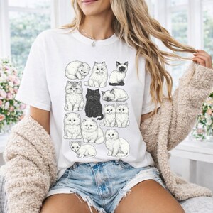 May include: White t-shirt featuring a collection of illustrated cats in various poses. The cats are depicted in black and white, with different breeds and fur patterns. The shirt is worn by a person in denim shorts and a beige cardigan.