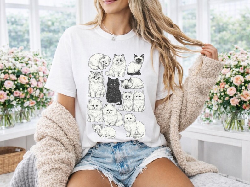 May include: White t-shirt featuring a collection of illustrated cats in various poses. The cats are depicted in black and white, with different breeds and fur patterns. The shirt is worn by a person in denim shorts and a beige cardigan.