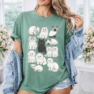 May include: A green t-shirt featuring a collection of illustrated cats in various poses. The cats are in shades of white, gray, and black. The shirt is a comfortable, casual style.