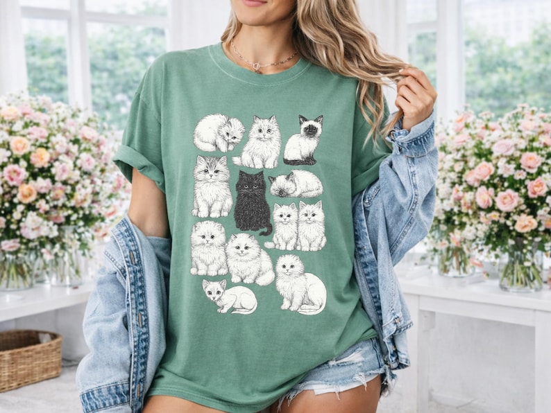 May include: A green t-shirt featuring a collection of illustrated cats in various poses. The cats are in shades of white, gray, and black. The shirt is a comfortable, casual style.