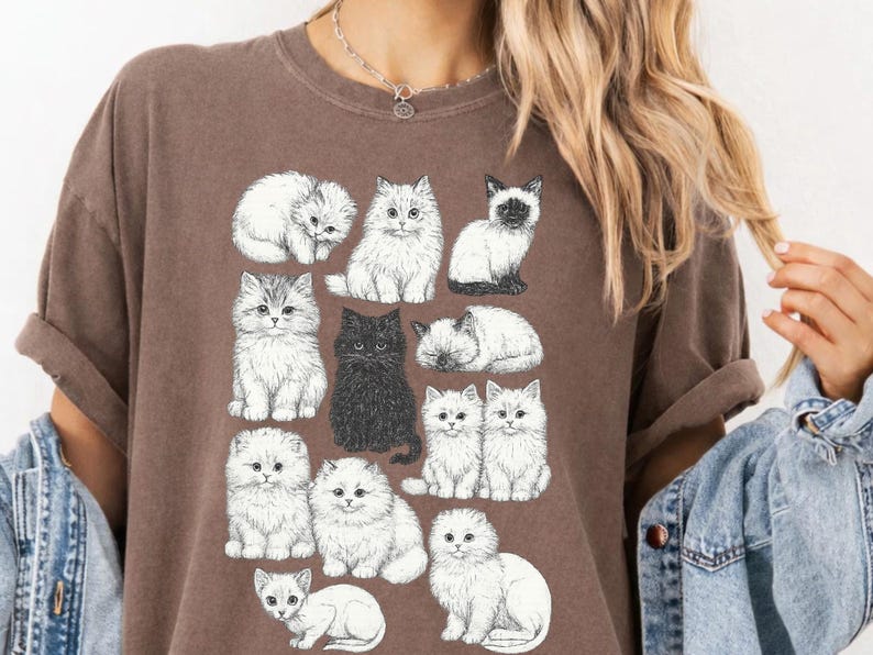 May include: Brown t-shirt featuring a collection of illustrated cats in various poses. The cats are primarily white, with one black cat and a Siamese cat. The design includes fluffy kittens, creating a whimsical and charming aesthetic.