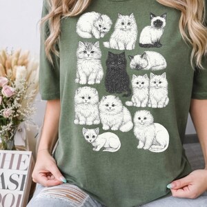 May include: Olive green t-shirt featuring a collection of illustrated cats in various poses. The cats are depicted in shades of white, gray, and black, with detailed fur and expressive faces. A charming and whimsical design for cat lovers.