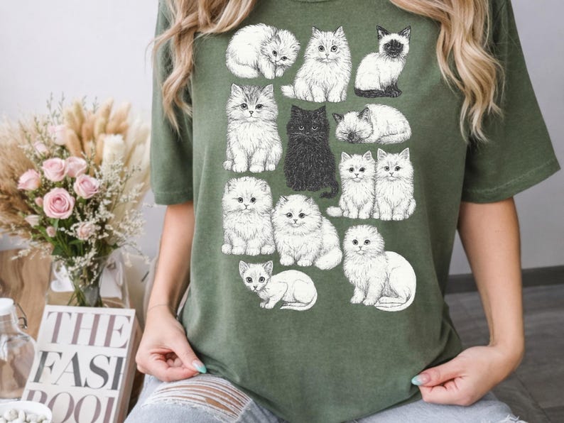 May include: Olive green t-shirt featuring a collection of illustrated cats in various poses. The cats are depicted in shades of white, gray, and black, with detailed fur and expressive faces. A charming and whimsical design for cat lovers.