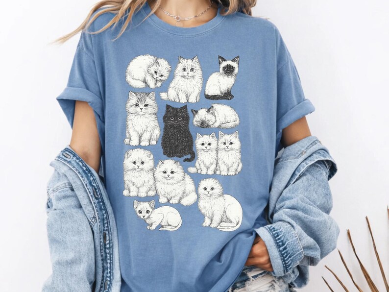 May include: A light blue t-shirt with a pattern of illustrated cats in different poses. The cats are in black and white, with one black cat. The shirt is a casual style, suitable for everyday wear.