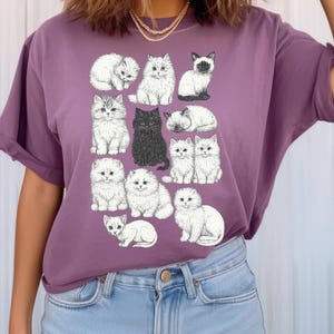 May include: Purple t-shirt featuring a collection of illustrated cats in various poses. The cats are primarily white with some black and grey accents. The design includes a black cat and a Siamese cat. The shirt is a solid purple color.