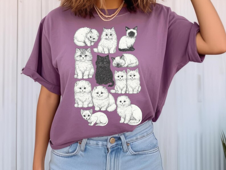 May include: Purple t-shirt featuring a collection of illustrated cats in various poses. The cats are primarily white with some black and grey accents. The design includes a black cat and a Siamese cat. The shirt is a solid purple color.