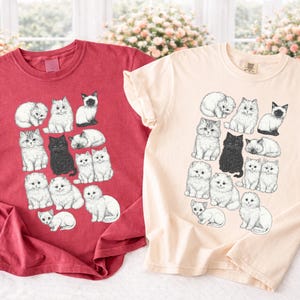 May include: Two short-sleeved t-shirts, one red and one cream, each featuring a repeating pattern of illustrated cats in various poses. The cats are in black and white, with some in a sitting or sleeping position.
