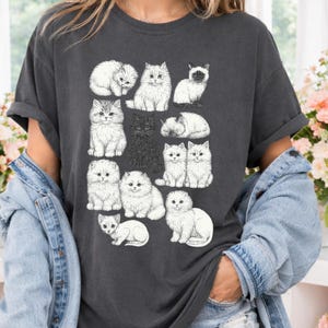 May include: Dark gray t-shirt featuring a collection of illustrated cats in various poses. The cats are depicted in a detailed, sketch-like style, with varying fur textures and expressions. The design includes both white and black cats.