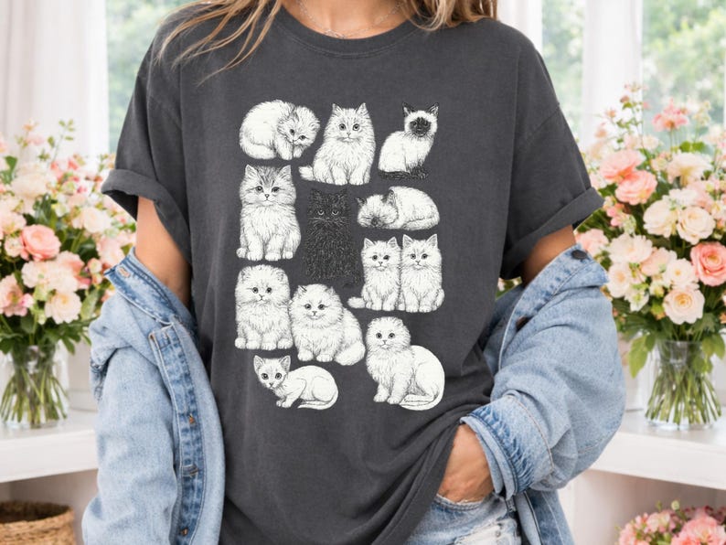 May include: Dark gray t-shirt featuring a collection of illustrated cats in various poses. The cats are depicted in a detailed, sketch-like style, with varying fur textures and expressions. The design includes both white and black cats.