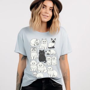 May include: Light blue t-shirt featuring a collection of illustrated cats in various poses. The cats are depicted in a detailed, black and white style, with one solid black cat. The shirt is a crew neck style.