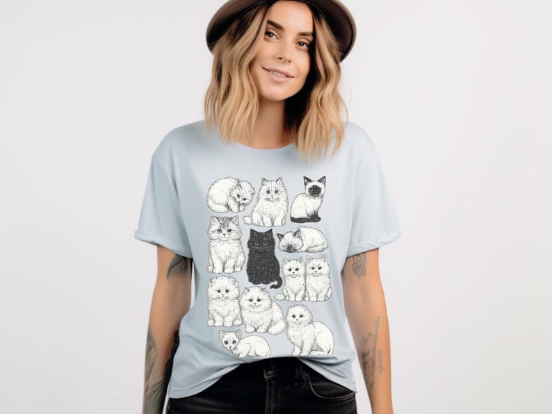 May include: Light blue t-shirt featuring a collection of illustrated cats in various poses. The cats are depicted in a detailed, black and white style, with one solid black cat. The shirt is a crew neck style.