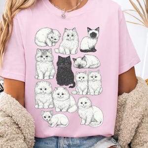 May include: Pink t-shirt featuring a collection of illustrated cats in various poses. The cats are depicted in a detailed, black and white style, with one black cat standing out. The shirt is a soft pink color.