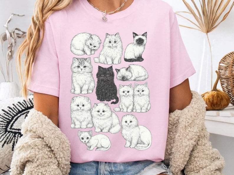 May include: Pink t-shirt featuring a collection of illustrated cats in various poses. The cats are depicted in a detailed, black and white style, with one black cat standing out. The shirt is a soft pink color.