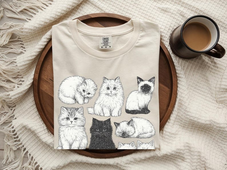 May include: A cream-coloured t-shirt with a detailed illustration of various cat breeds. The black and white drawings include fluffy Persians, a Siamese, and a black cat. The shirt is on a wooden tray with a mug of coffee.