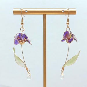 Puede incluir: Un par de pendientes colgantes con dijes de flores de iris moradas y detalles de alambre en tono dorado. Cada pendiente tiene un tallo largo y fino en tono dorado con una hoja verde pálido y una pequeña cuenta transparente en forma de gota.
