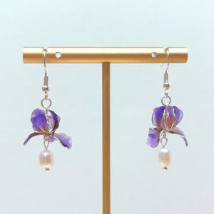 May include: A pair of drop earrings featuring translucent purple iris flower charms with silver-toned wire edges, each accented with a single dangling white freshwater pearl, suspended from silver-toned metal ear hooks.