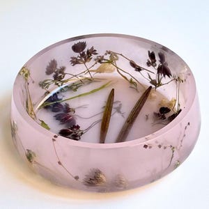 May include: A round, translucent light pink resin bowl featuring embedded dried dark purple flowers, green stems, and two elongated brown seed pods. The bowl has a smooth, polished finish with the botanical elements visible throughout the material.