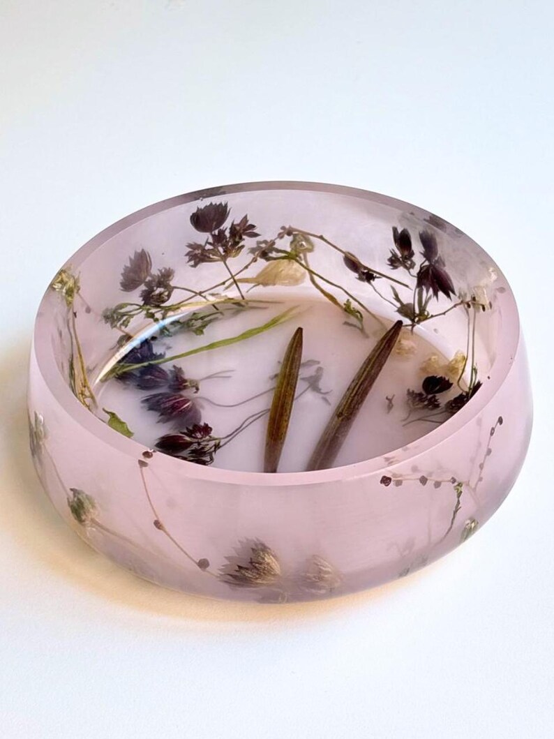 May include: A round, translucent light pink resin bowl featuring embedded dried dark purple flowers, green stems, and two elongated brown seed pods. The bowl has a smooth, polished finish with the botanical elements visible throughout the material.