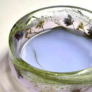 Può includere: Una ciotola circolare in resina trasparente con elementi botanici essiccati incorporati, tra cui erbe verdi e piccoli fiori bianchi e viola. La base della ciotola presenta un colore lavanda chiaro solido e opaco, con lati trasparenti.