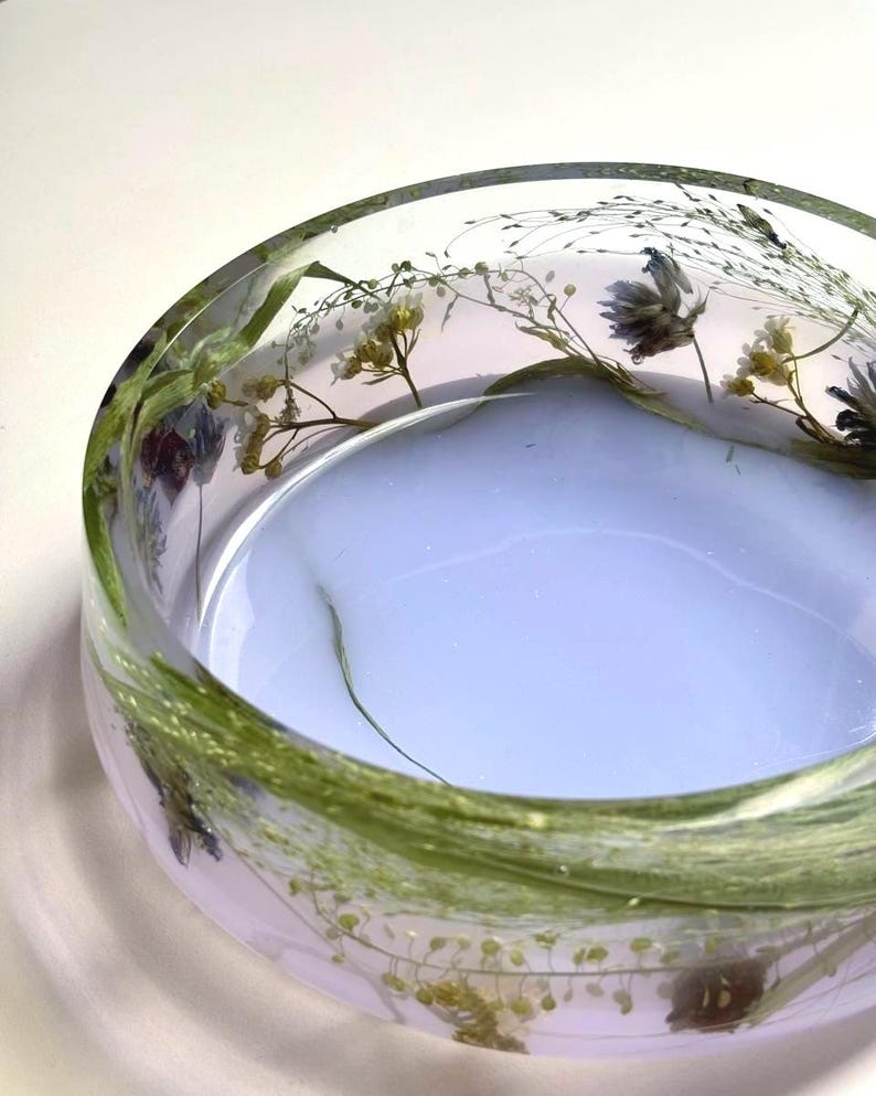 May include: A circular, clear resin bowl featuring embedded dried botanical elements, including green grasses and small white and purple flowers. The base of the bowl has a solid, opaque light lavender color, while the sides remain transparent.