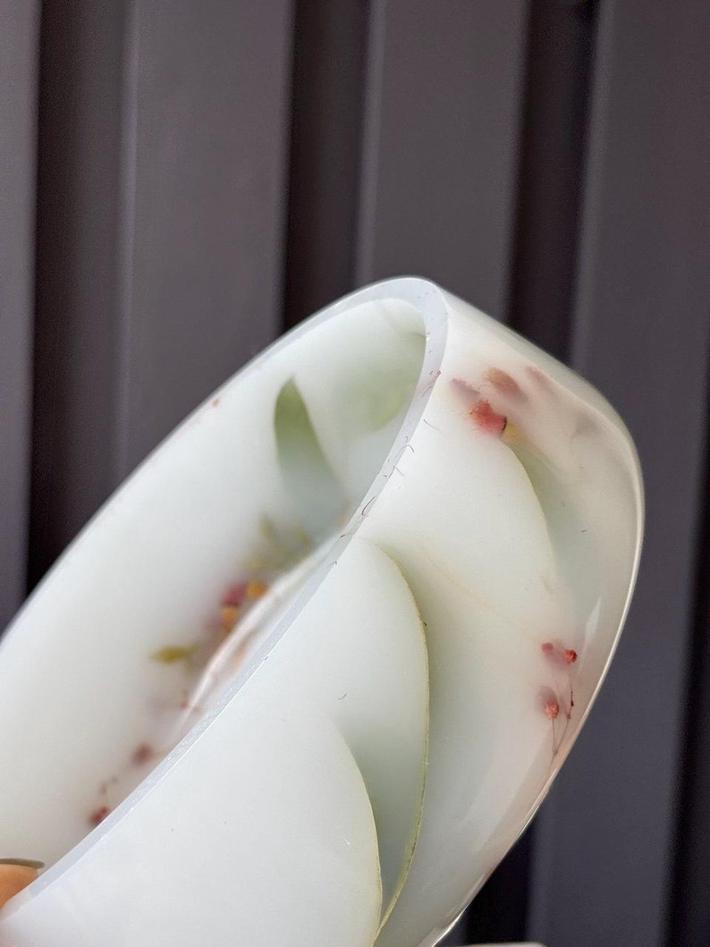 May include: A translucent white resin bangle bracelet featuring embedded dried green leaves and small pink flower buds. The circular accessory has a smooth, polished finish and a rounded profile, showcasing the delicate botanical elements suspended within.