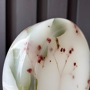 May include: A smooth, rounded, translucent white resin object featuring embedded dried green leaves and small red flowers. The surface is polished and glossy, showcasing the botanical elements suspended within the clear, milky material.