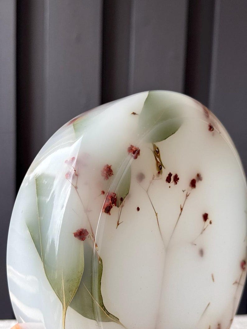 May include: A smooth, rounded, translucent white resin object featuring embedded dried green leaves and small red flowers. The surface is polished and glossy, showcasing the botanical elements suspended within the clear, milky material.