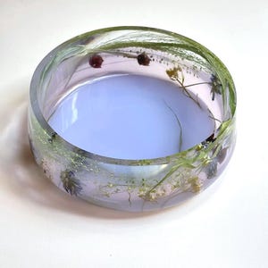 May include: A round, clear resin bowl featuring embedded dried green grasses, small white flowers, and tiny dark red buds. The base of the bowl has a solid, opaque light lavender tint, while the sides remain transparent, showcasing the botanical elements.