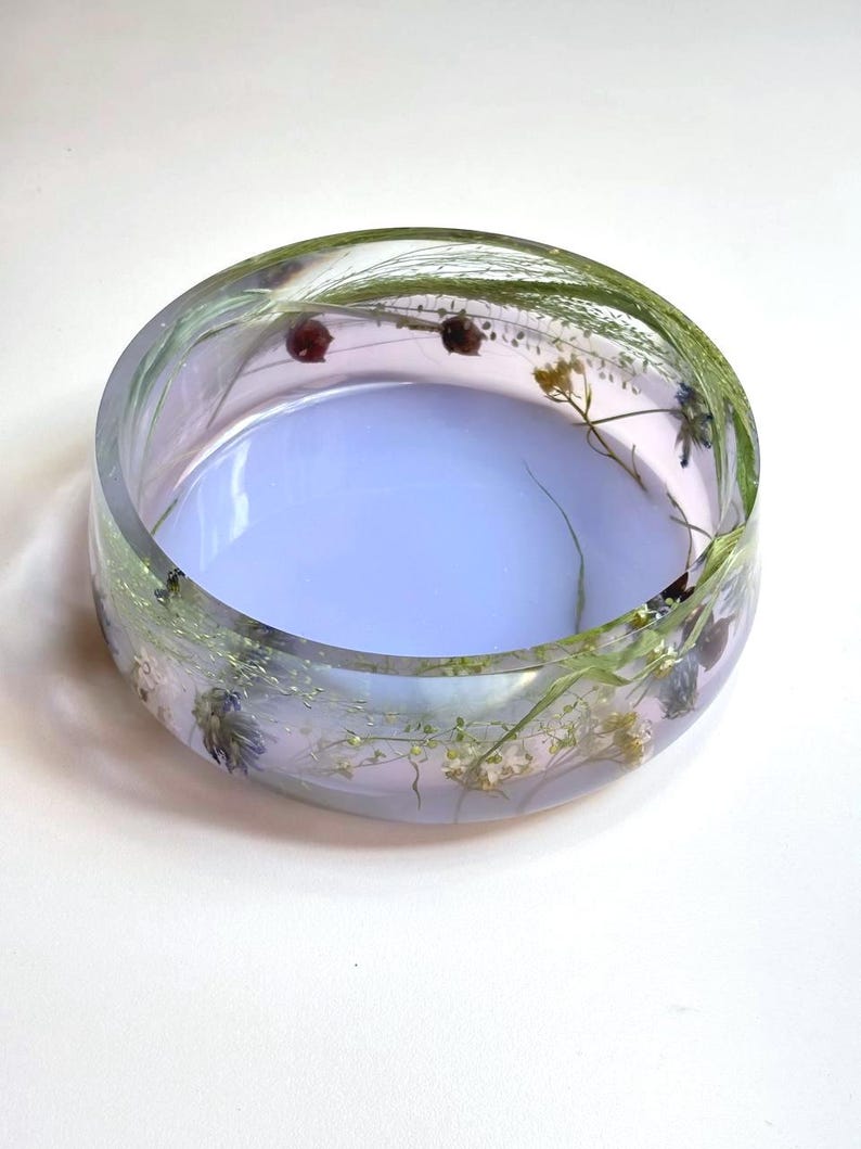 May include: A round, clear resin bowl featuring embedded dried green grasses, small white flowers, and tiny dark red buds. The base of the bowl has a solid, opaque light lavender tint, while the sides remain transparent, showcasing the botanical elements.