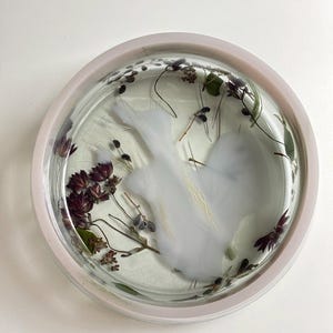 May include: A circular clear resin tray featuring embedded dried dark purple flowers, green foliage, and a central opaque white swirl pattern. The tray has a smooth, glossy finish and a raised rim, showcasing a botanical arrangement preserved within the transparent material.
