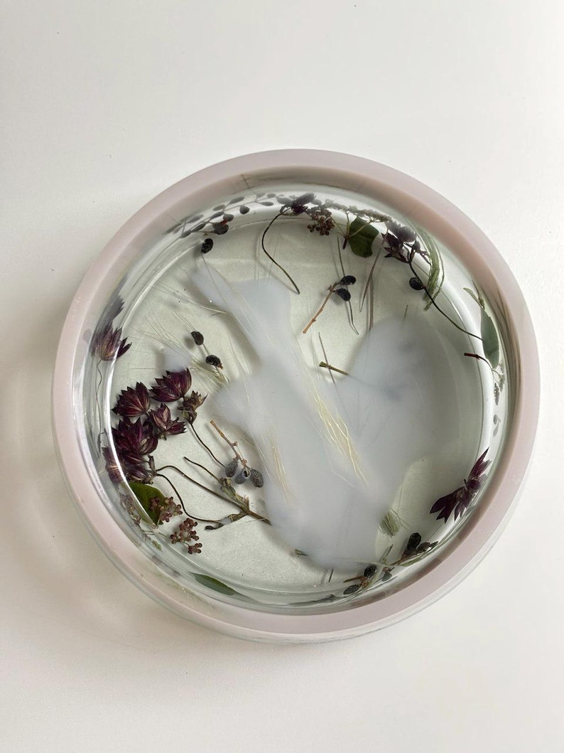 May include: A circular clear resin tray featuring embedded dried dark purple flowers, green foliage, and a central opaque white swirl pattern. The tray has a smooth, glossy finish and a raised rim, showcasing a botanical arrangement preserved within the transparent material.