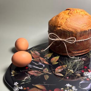 Puede incluir: Un panettone horneado, atado con hilo, se encuentra en una bandeja negra decorada con hojas y flores. Dos huevos marrones también están en la bandeja. La bandeja tiene un acabado brillante.
