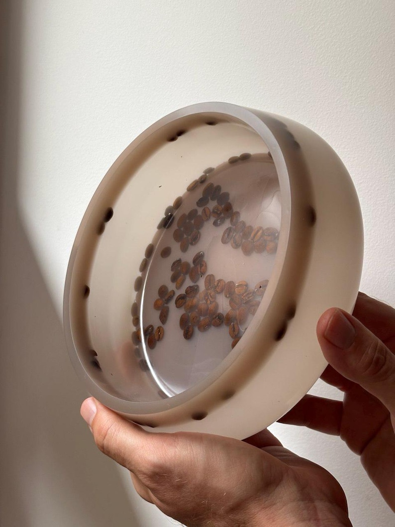 May include: A circular, translucent off-white resin bowl featuring embedded dark brown roasted coffee beans. The beans are arranged in a scattered pattern within the base and along the interior side walls of the bowl, creating a visible contrast.