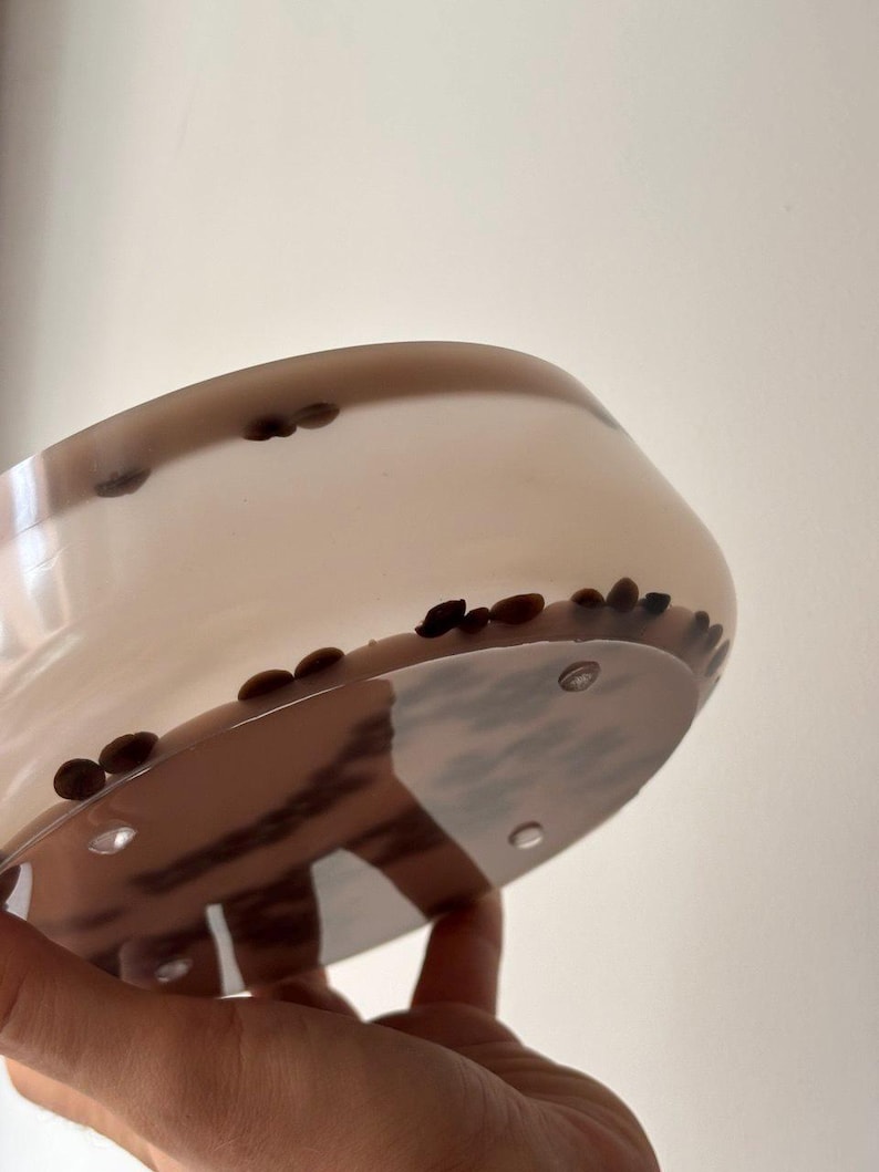 May include: A round, translucent, light beige bowl with a smooth, glossy finish. The bowl is embedded with numerous dark brown, oval-shaped coffee beans distributed throughout the material, visible through the semi-transparent walls.