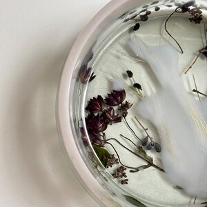 May include: A circular tray with a pale pink rim, featuring a clear resin base embedded with dried dark purple flowers, delicate green stems, and a swirling opaque white pigment effect that creates a cloud-like pattern within the material.