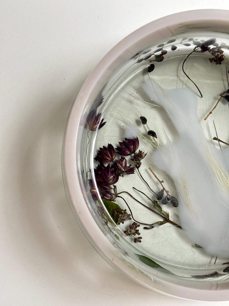 May include: A circular tray with a pale pink rim, featuring a clear resin base embedded with dried dark purple flowers, delicate green stems, and a swirling opaque white pigment effect that creates a cloud-like pattern within the material.