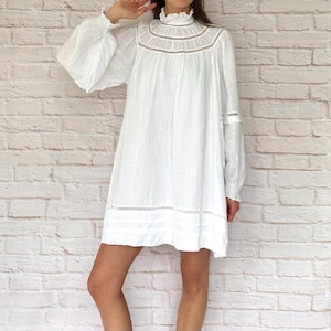 Pode incluir: Vestido de linho branco com mangas compridas e volumosas e gola alta com babados. O vestido apresenta detalhes delicados de renda ao redor do pescoço, mangas e bainha. O vestido é na altura do joelho e tem uma silhueta fluida.