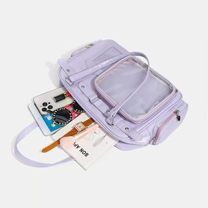May include: A lavender handbag with a clear, rectangular pocket on the front. The bag is open, revealing a smartphone, sunglasses, a watch, and a small notebook with the text "BON APP!". The bag has a shoulder strap and a zipper closure.