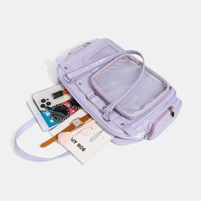 May include: A lavender handbag with a clear, rectangular pocket on the front. The bag is open, revealing a smartphone, sunglasses, a watch, and a small notebook with the text "BON APP!". The bag has a shoulder strap and a zipper closure.