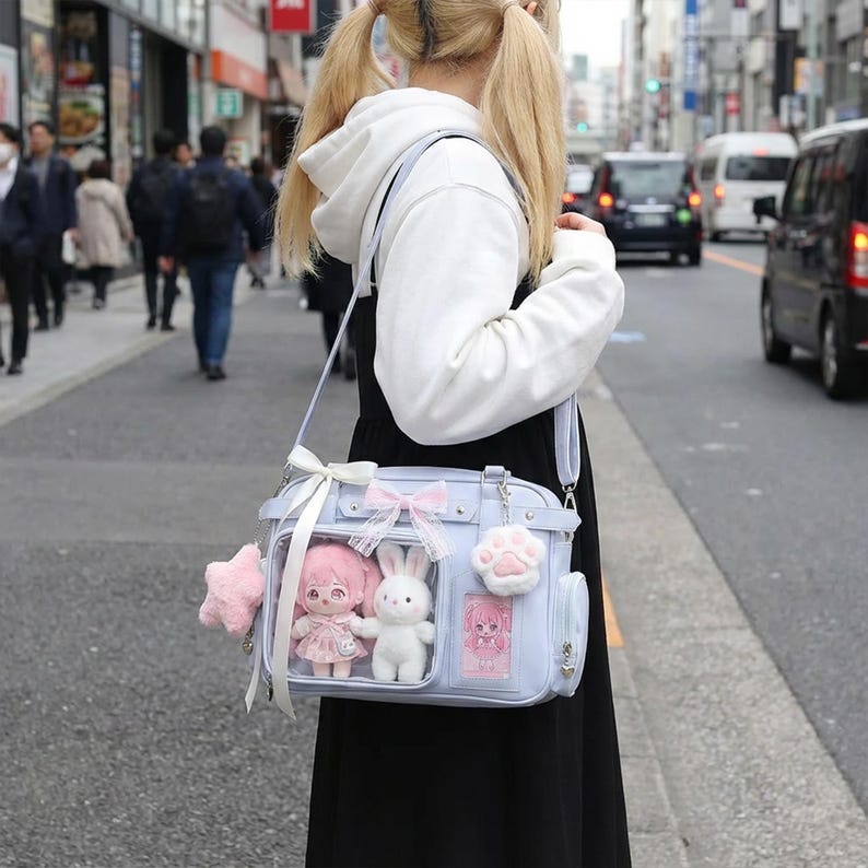May include: A light blue shoulder bag with a clear front panel, decorated with pink bows and star-shaped and paw-shaped charms. The bag contains a pink doll and a white bunny plushie. The bag is carried on a street.