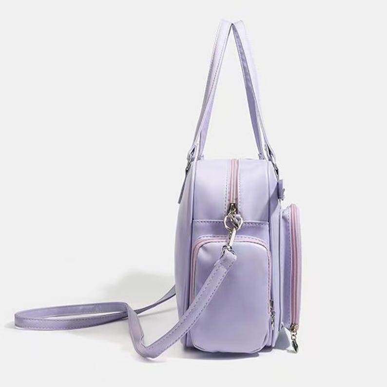 May include: A lavender handbag with a shoulder strap and two handles. The bag has a rectangular shape with a zippered main compartment and two exterior pockets. The zippers and trim are a lighter shade of pink.