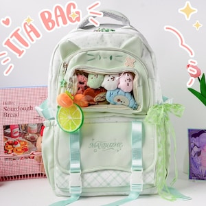 Könnte beinhalten: Ein hellgrüner Rucksack mit einem durchsichtigen Sichtfenster, das mit kleinen Plüschtieren gefüllt ist, Katzenohren-Akzenten und dem Text MANBUZHE auf der Vordertasche. Der Rucksack hat zwei Schnallenriemen, einen Limettenscheiben-Anhänger und Spitzenbesatz.