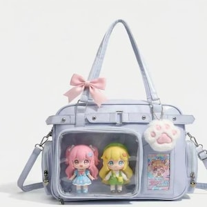 May include: A light blue handbag with a pink bow, featuring two anime-style dolls in a clear compartment. The bag has a shoulder strap and a paw-shaped charm. The dolls are wearing dresses.