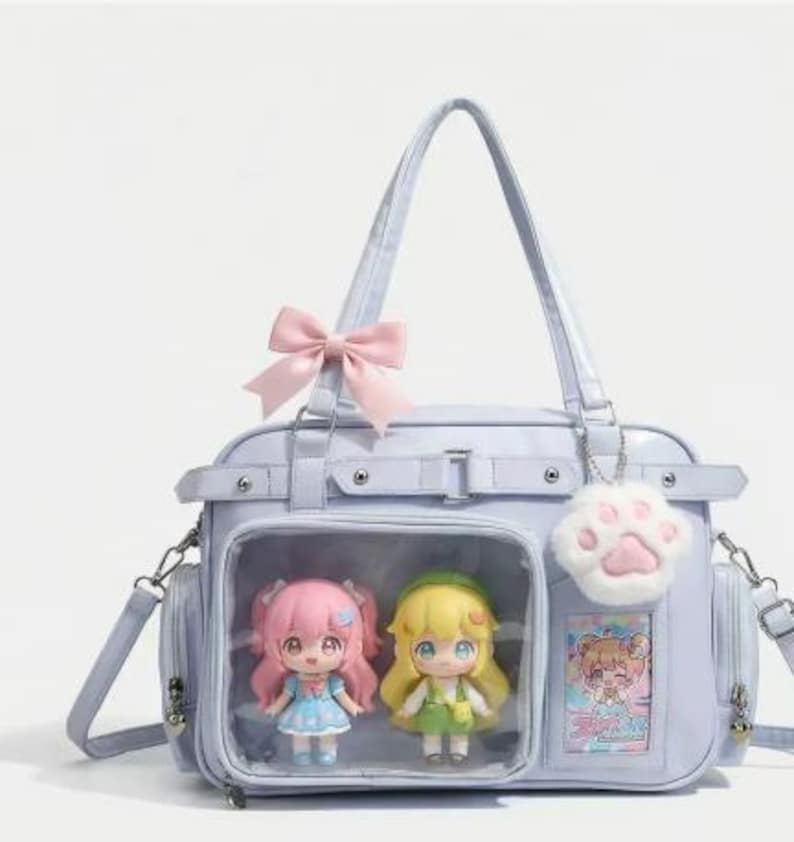 May include: A light blue handbag with a pink bow, featuring two anime-style dolls in a clear compartment. The bag has a shoulder strap and a paw-shaped charm. The dolls are wearing dresses.